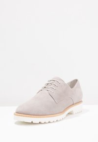 Scarpa stringata in suede grigio chiaro con punta arrotondata, suola in gomma bianca e accento beige lungo il bordo della suola. Texture liscia su tutta la superficie.