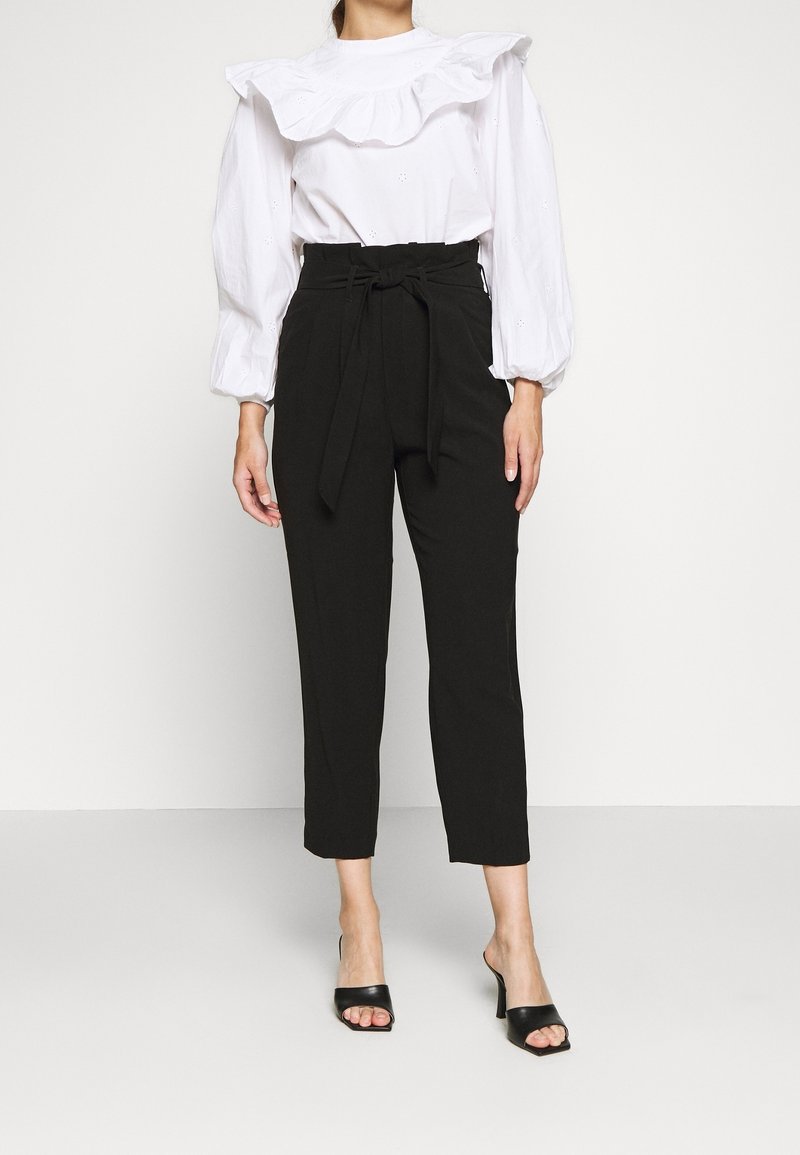 Femme portant un chemisier blanc à volants avec manches bouffantes, un pantalon noir taille haute fuselé avec une ceinture à la taille, et des talons ouverts noirs.