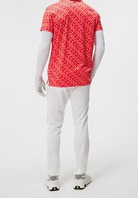 Camiseta de manga corta con patrón rojo y diseño geométrico, combinada con pantalones blancos y zapatos deportivos con acentos grises, mostrando un guante en una mano.