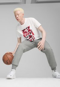 Hvid T-shirt med rødt grafisk tryk, grå sweatpants og hvide sneakers. Holder en brun basketball og poserer i en lav stilling.