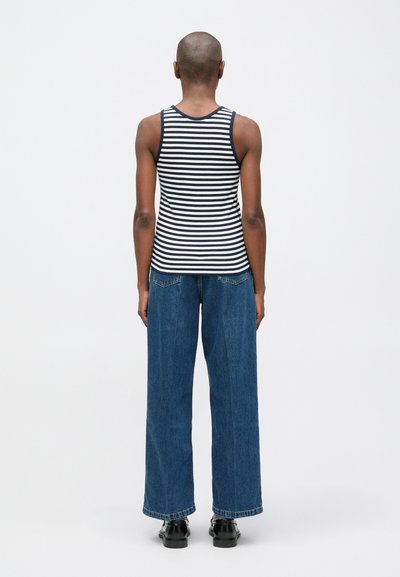 Cilvēks ar noskūtu galvu, tērpies tumši zilo un baltu svītrainu bezpiedurkņu krekšu, platām zilo džinsu biksēm un melnām kurpēm, stāv ar muguru vērsts.