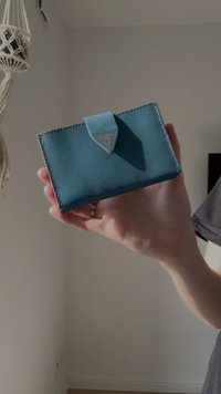 Turquoise cardholder made of textured material, featuring a triangular accent and a folded flap for closure. Handheld against a neutral background.
