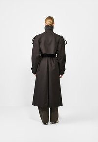 Osoba z blond ciasno związanym kokiem, ubrana w długi ciemny płaszcz z wysokim kołnierzem, podkreśloną talią i szerokimi spodniami, stojąca na tle jednolitego białego tła.
