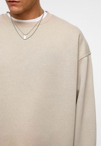 Beige sweatshirt met een ronde hals en verlaagde schouders, gemaakt van zachte stof. Twee zilveren kettingen zijn over een witte shirt gelaagd.