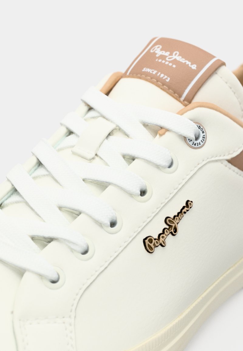 Zapatilla blanca de piel con un acabado suave, que cuenta con cordones blancos, un acento marrón claro en la lengüeta y un logo dorado.