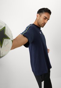 T-shirt en coton bleu marine à manches courtes, avec un logo sur la poitrine et des détails de bandes contrastantes le long des épaules.