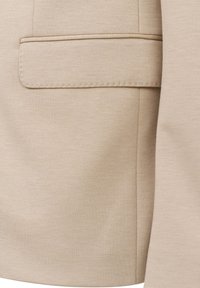 Beige Blazerstoff mit glatter Textur, einem einzelnen Fach, subtilen Nähdetails und einer strukturierten Form.