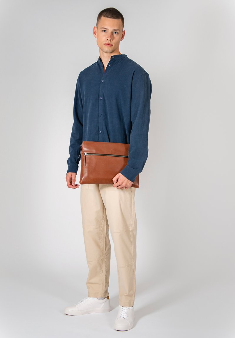 Braune Lederlaptoptasche mit einem Reißverschlussfach, gehalten von einer Person in einem blauen Hemd und beigen Hosen, mit weißen Turnschuhen.