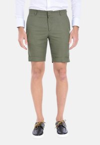 Pantaloni corto verde khaki in un tessuto leggero, con una vestibilità dritta e orlo arrotolato, abbinati a scarpe nere con lacci e suole bianche.