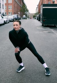 Atletska oblačila: črna jakna z zadrgo in oprijete črne legice. Bele anklet nogavice in črne športne čevlje s srebrnimi detajli. Urbani ulični ambient.