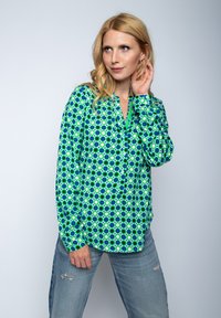 Bluzka z długim rękawem w intensywnym zielonym i niebieskim okrągłym wzorze, z dekoltem w kształcie litery V i detalem w postaci guzików, wykonana z gładkiego materiału. Noszona z przetartymi dżinsami.