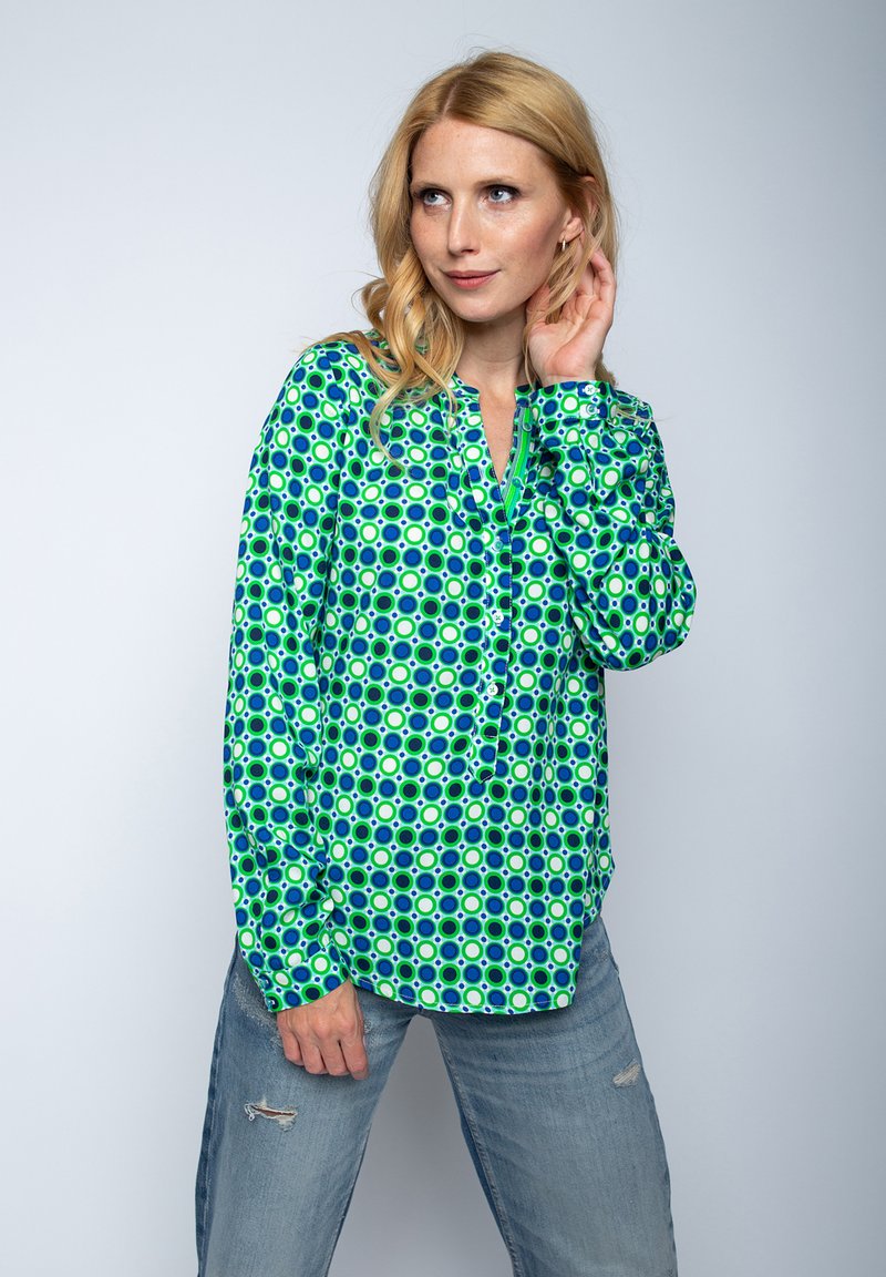 Bluzka z długim rękawem w intensywnym zielonym i niebieskim okrągłym wzorze, z dekoltem w kształcie litery V i detalem w postaci guzików, wykonana z gładkiego materiału. Noszona z przetartymi dżinsami.