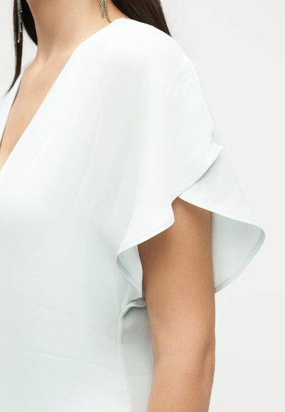 Γυναίκα που φοράει λευκό τοπ με φουσκωτά μανίκια και ντεκολτέ σε σχήμα V, με ακάλυπτους ώμους και πάνω από το χέρι, με μακριά σκούρα μαλλιά και μπλε σκουλαρίκια με χάντρες.