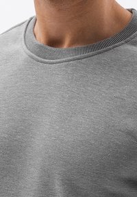 Primo piano di una persona che indossa un maglione grigio a girocollo, in cui si vedono il colletto e la parte superiore del petto.