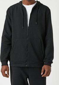 Sudadera con cremallera negra hecha de tela texturizada, con una capucha ajustada, dos bolsillos frontales y detalles de cordón. Diseño simple y moderno.