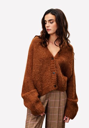 Cardigan de couleur rouille, surdimensionné et fabriqué dans un matériau duveteux, avec un col en V, de gros boutons et des poignets larges, associé à un pantalon à carreaux.