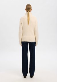 Pulover tricotat de culoare crem, cu guler înalt, tiv cu nervuri și mâneci lungi, asortat cu blugi negri din denim cu picioare drepte.