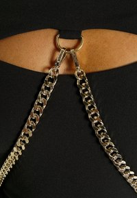 Des sangles en chaîne dorée fixées par des fermoirs à une petite bague dorée sur un vêtement découpé noir, laissant apparaître une peau nue en dessous.