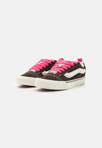 Bruna mockaskor med rosa snören, vita accenter och en texturerad gummisula. Har en låg design och en rosa dragflik vid hälen.