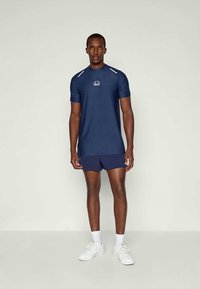 Marine atletisk skjorte med korte ermer, strukturert stoff, hvit logo på brystet, kombinert med matchende shorts og hvite joggesko.