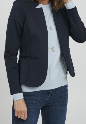 Femme portant un blazer bleu marine par-dessus un pull bleu clair avec une broderie florale et un jean bleu foncé, debout devant un fond uni.