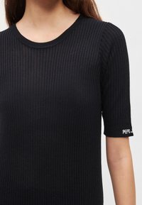 Μαύρο κοντομάνικο ριπ τοπ με στρογγυλή λαιμόκοψη. Διαθέτει ορατή λεπτομέρεια λογότυπου σε λευκό στο μανίκι. Λείο υφές.