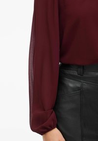Blusa trasparente bordeaux con maniche lunghe a sbuffo abbinata a una gonna nera in pelle aderente con cuciture dettagliate e tasca laterale.