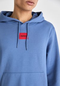 Felpa azzurra chiara con tasca a marsupio frontale, decorata con una patch rettangolare rossa su cui è scritto "HUGO" in testo nero. Realizzata in un tessuto morbido, con cordini.