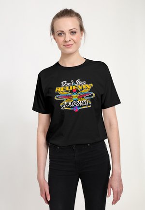 Frau trägt ein schwarzes T-Shirt mit buntem Grafikdesign und dem Text "Don't Stop Believin' Journey" vor einem weißen Hintergrund.
