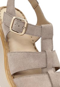 Sandalo in suede beige con tacco a zeppa, dotato di cinghie incrociate e dettaglio in metallo sulla cinghia alla caviglia. Soletta con superficie liscia.
