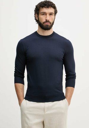 Uomo con capelli scuri e ricci e barba, indossa una camicia aderente blu scuro a maniche lunghe e pantaloni beige chiaro, in piedi con le mani in tasca.