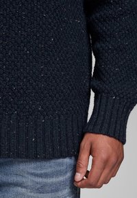 Marineblauwe gebreide trui met textuurpatroon op het lichaam en geribde manchetten, met kleurvlekken verspreid over het geheel en een ontspannen pasvorm.