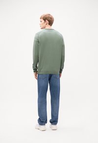 Long-sleeve green shirt with a ribbed crew neckline, paired with blue denim jeans. White sneakers visible at the bottom. Simple, modern design.