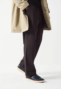 Pantaloni marroni scuri con una vestibilità rilassata, abbinati a sneakers in pelle blu navy con suola crema, e un cappotto beige indossato in posizione eretta.