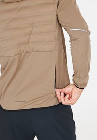 Leichte, beige Jacke mit gepolstertem oberen Teil, elastischen Bündchen und seitlichen Reißverschlusstaschen. Verfügt über reflektierende Akzente und eine glatte Textur.