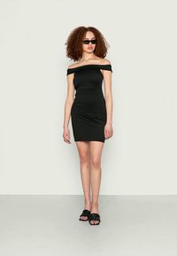 Vestido justo preto com decote ombro a ombro e design ajustado. Modelo usa óculos de sol pretos e sandálias rasteiras texturizadas pretas.