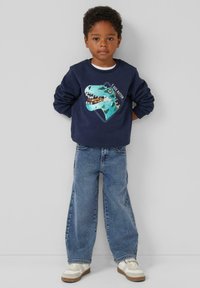 Marineblauer Sweatshirt mit einem türkisfarbenen Dinosaurier-Motiv, kombiniert mit lässigen Jeans im Loose-Fit und weißen Sneakers. Lässiges und verspieltes Outfit.