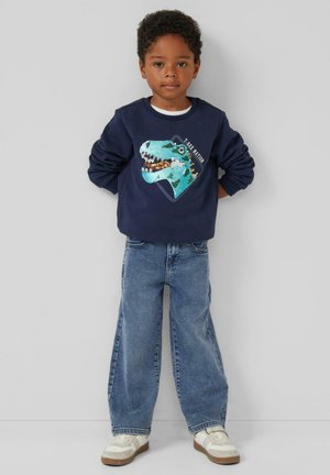 Marineblauwe sweatshirt met een turquoise dinosaurustekening, gecombineerd met losvallende spijkerbroeken en witte sneakers. Casual en speelse outfit.