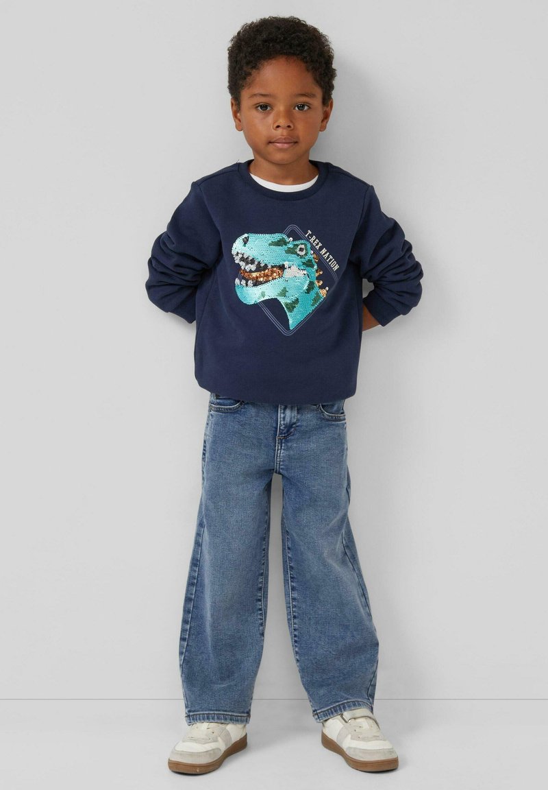 Marineblauer Sweatshirt mit einem türkisfarbenen Dinosaurier-Motiv, kombiniert mit lässigen Jeans im Loose-Fit und weißen Sneakers. Lässiges und verspieltes Outfit.