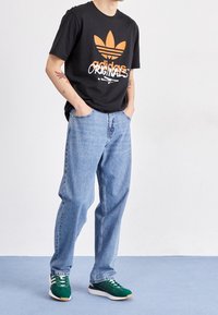 Černé bavlněné tričko s oranžovým logem Adidas, spárované se světle modrými džíny a zelenými teniskami s bílými detaily a tkaničkami.