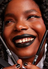 Svart flytende eyeliner og svart leppestift holdes i hånden. Subjektet har flettehår, en glatt hudtone, og smiler bredt.