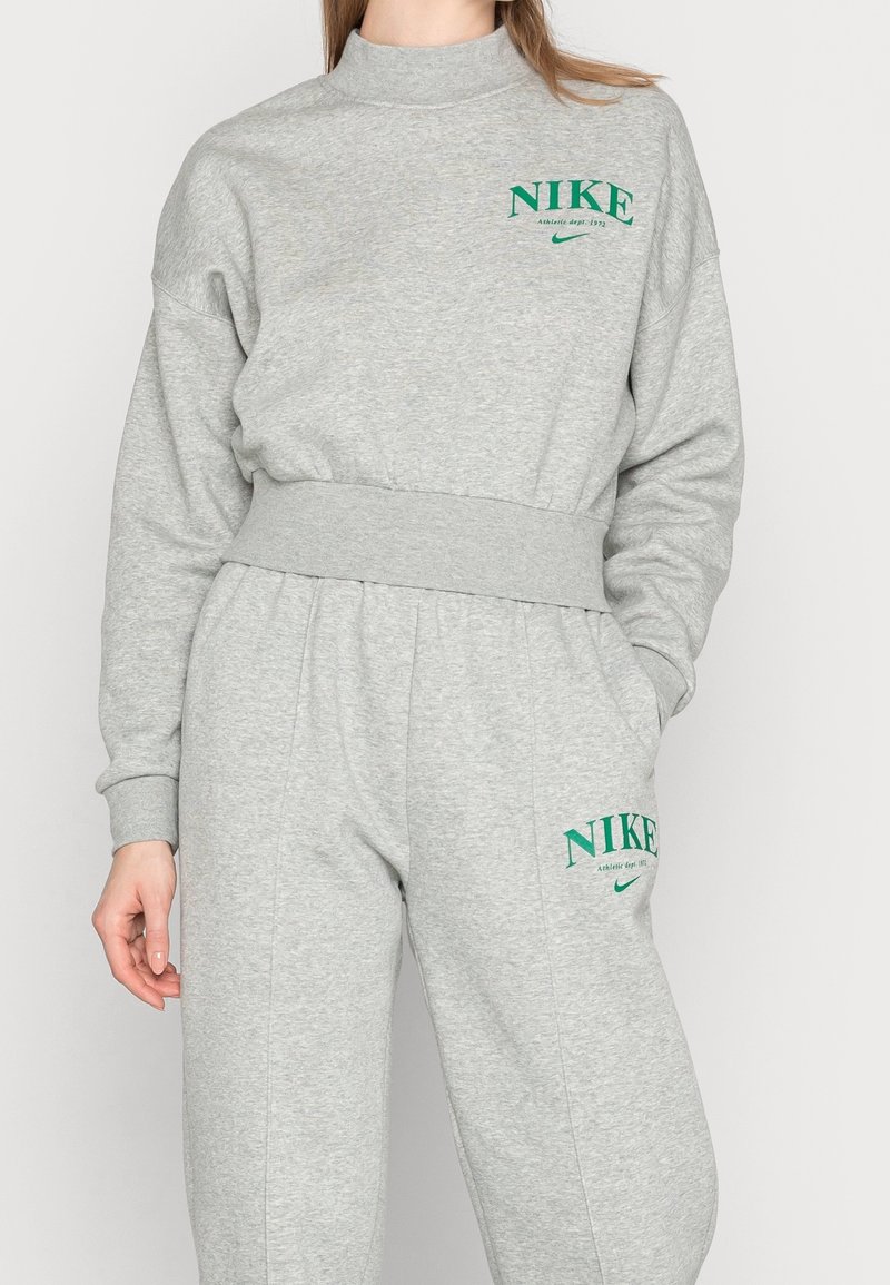 Kvinna iklädd grå Nike-tröja och matchande sweatpants med grön Nike-logotyp och text, stående mot en enkel bakgrund.