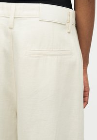 Κοντινή λήψη ενός ατόμου που φοράει εκρού παντελόνια με πίσω τσέπη με φάσα και θηλιά ζώνης, το χέρι ακουμπάει στο πλάι.