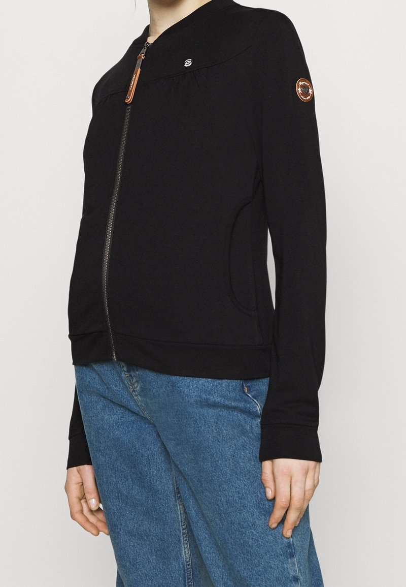 Personne portant une veste noire zippée avec un patch de logo rond sur la manche et un jean bleu, sur un fond uni.