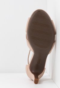 Nøgen højhælet sandal med en glat syntetisk overdel, en slank rem og en let tekstureret brun gummisål. Har en skulptureret hæl.