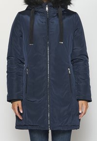 Abrigo acolchado azul marino con capucha de piel sintética, cremallera frontal y dos bolsillos laterales con cremalleras. Mangas largas con puños elásticos.