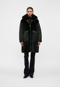 Palton din blană artificială neagră cu guler înalt, închidere cu fermoar, buzunare mari și mâneci contrastante, purtat peste pantaloni negri. Textură netedă și design călduros.