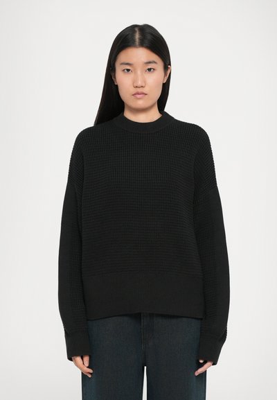 Suéter negro de punto con cuello redondo, corte holgado y patrón texturizado. Las mangas largas llegan más allá de las muñecas, combinadas con pantalones de mezclilla oscuros.