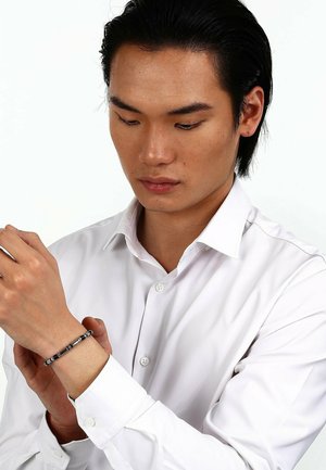 Schwarzes und silbernes Perlenarmband am Handgelenk, mit runden Perlen und einem strukturierten Mittelstück, getragen über einem weißen Button-up-Hemd.