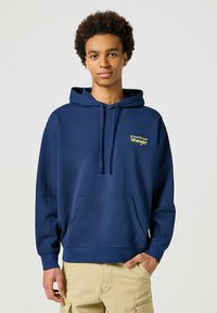 Granatowy hoodie z kieszenią kangurką, kapturem ze sznurkiem i haftowanym żółtym logo "Wrangler" na lewej piersi. Miękka faktura materiału.